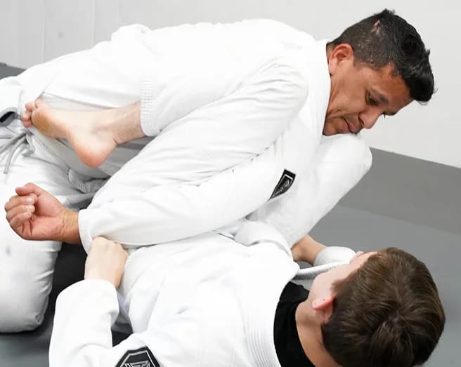 A coach in a white gi works a guard position with a student on the mat.